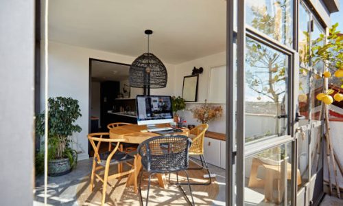 Sunroom1 View through open sliding glass door of sunny workspace adjoining kitchen with round table, chairs, pendant light, and desktop PC.